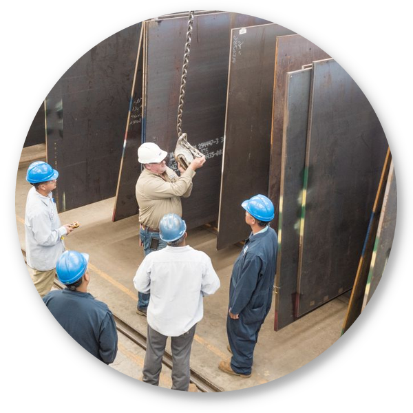 Employees in hard hats gathered around a supervisor demonstrating steel plate handling in a warehouse.
